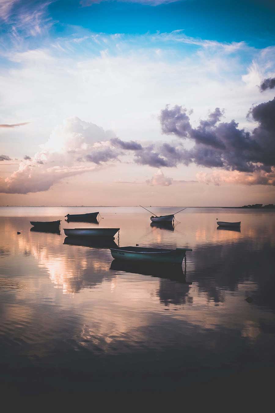 This is an image of 6 boats floating on the calm ocean with the sun slowly setting in the background